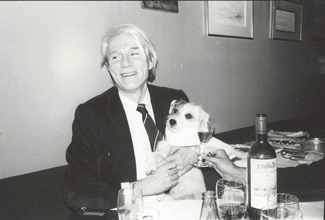 An Andy Warhol-esque man in a suit holding a dog at a table.