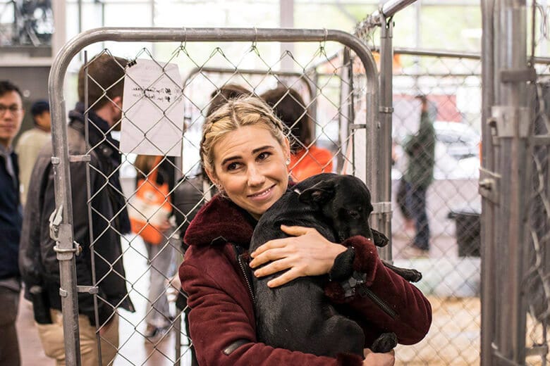 A woman holding a black dog at a mega-adoption event in front of a fence.