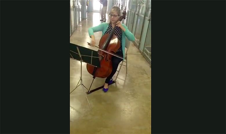 A woman playing classical music on a cello in a hallway while dogs listen attentively.