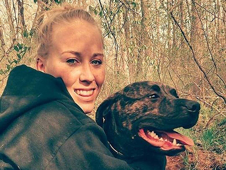 A woman in a hoodie walks apprehensively through the woods, tightly clutching onto her dog.