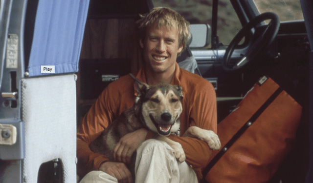 A man sitting in the back seat of a van with a dog, paying tribute to dogs.