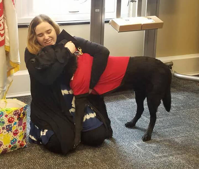 During a retiring ceremony, a woman affectionately hugs her service dog, a black Labrador Retriever, in an office.