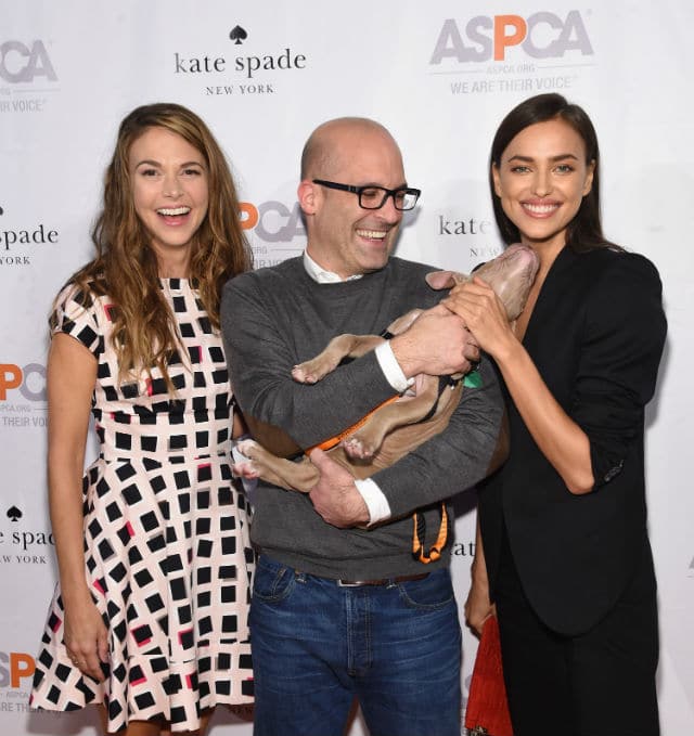 Three young friends posing with a dog on a red carpet at a benefit event.