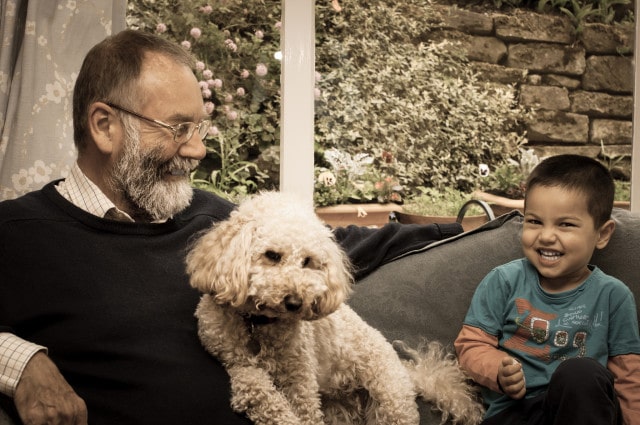 An older man and a young boy sitting on a couch with a poodle, enjoying meals together.