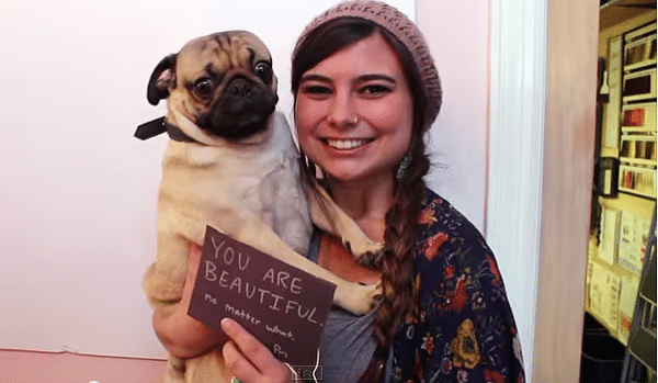 A woman holding Doug the Pug with a sign that says you are beautiful.