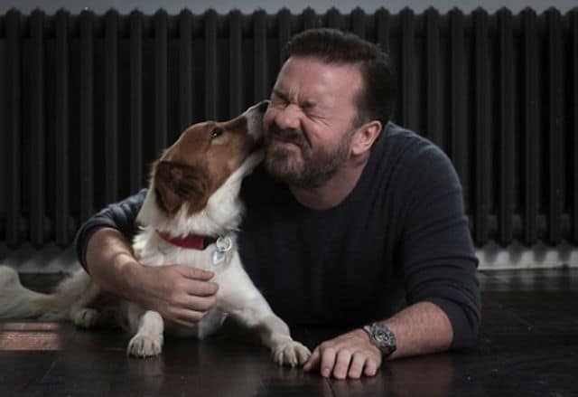 A man, resembling Ricky Gervais, lovingly kissing a dog on the floor.