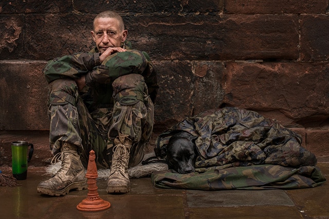 A man in camouflage sits on the ground next to a chess set, seemingly undetected.