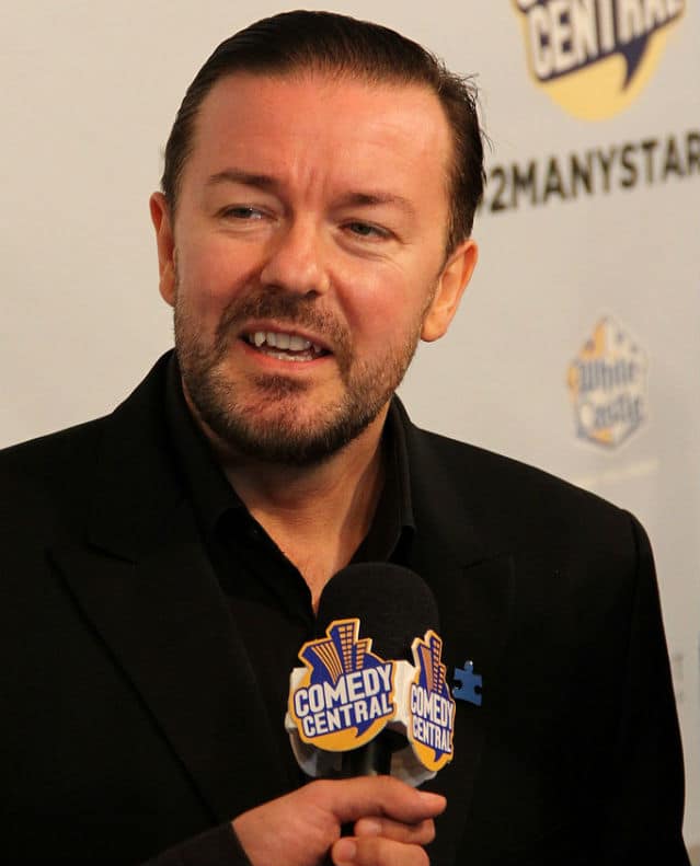 Ricky Gervais, a man in a suit, is holding a microphone.