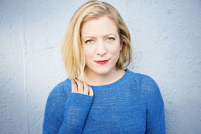 A woman in a blue sweater posing against a wall with an authoritative and confident stance (ACC).