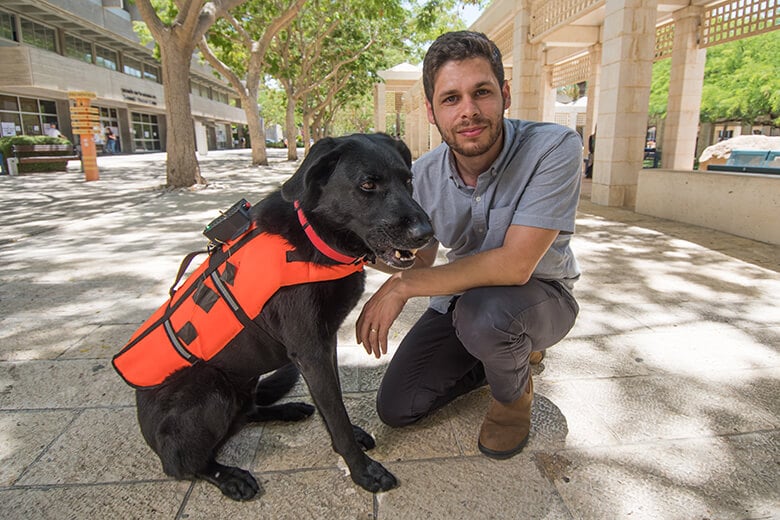 A man kneeling next to a black dog wearing a vibrating vest, teaching it commands without words.