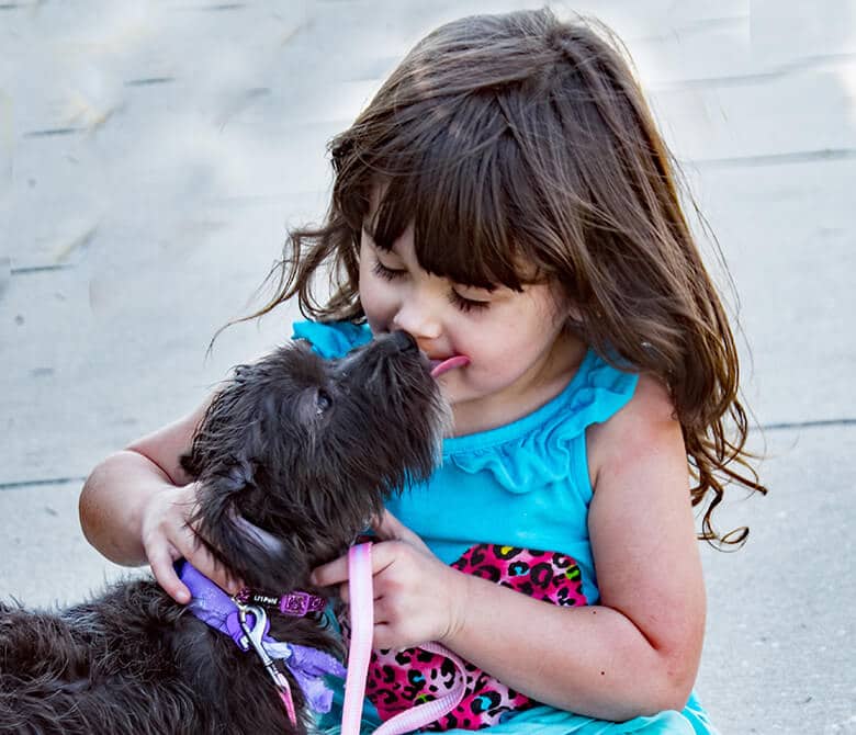 A little girl gently kissing a black dog. This Dogs Life