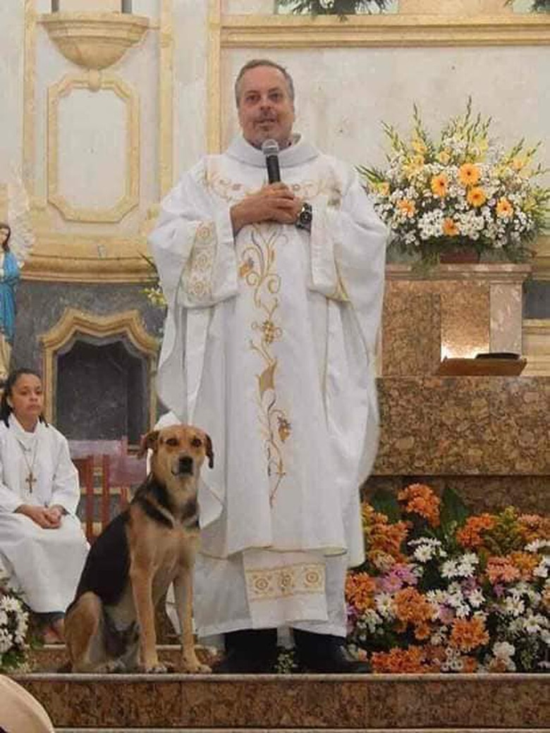 A priest in a white robe standing next to an adopted dog outside a church.