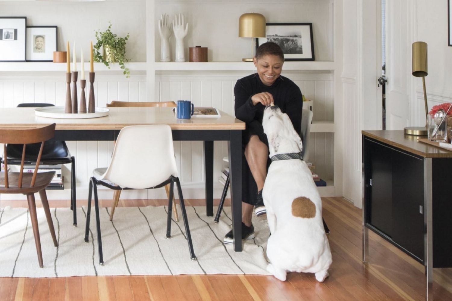 A woman is petting her dog in a dining room, showcasing products from Black-Owned Dog Businesses.