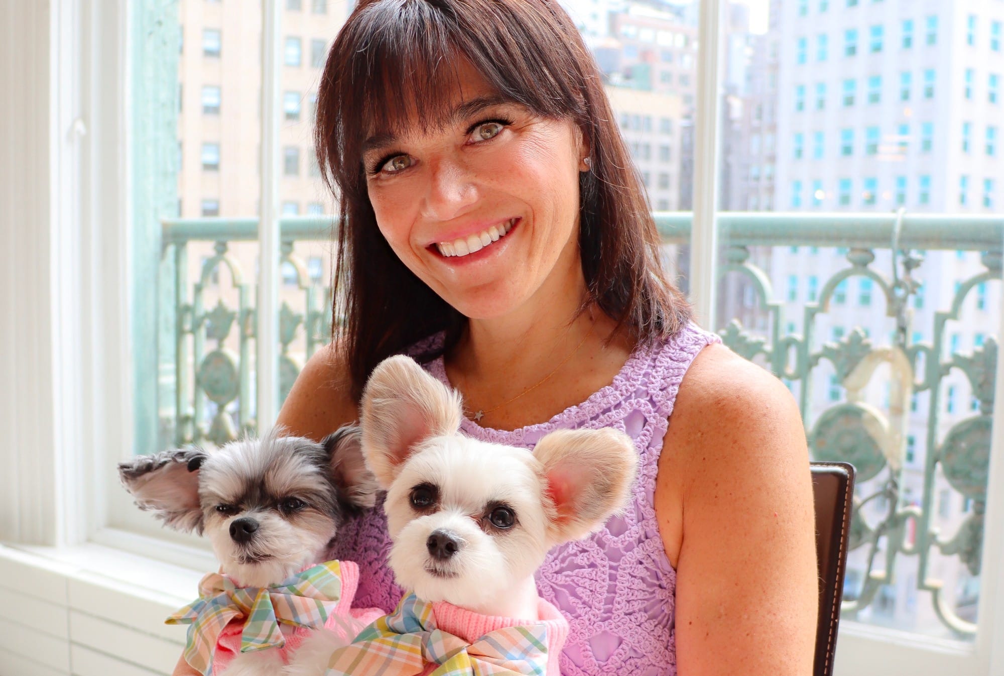 A woman holding two small pups in a pink bow tie celebrates International Dog Day.