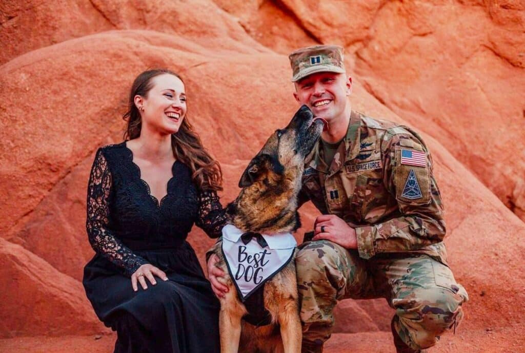 A military couple poses with their rescue dog in front of red rocks.