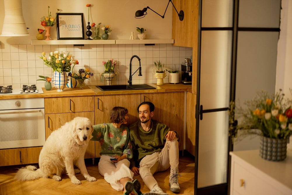 A man and woman sitting on the floor in a kitchen with their dog.